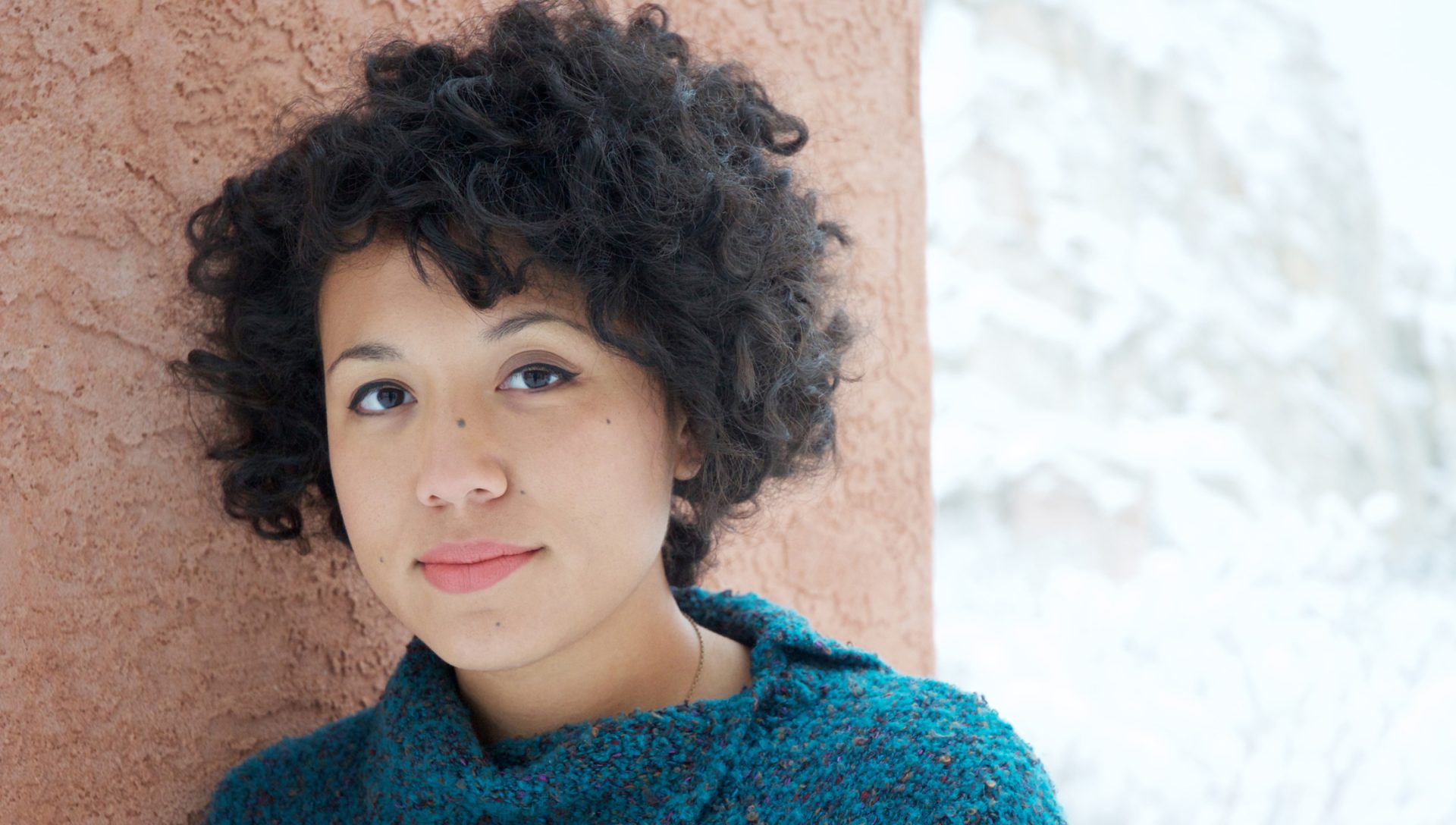 a women with short curly hair is smiling at the camera, she is leaning against a wall.
