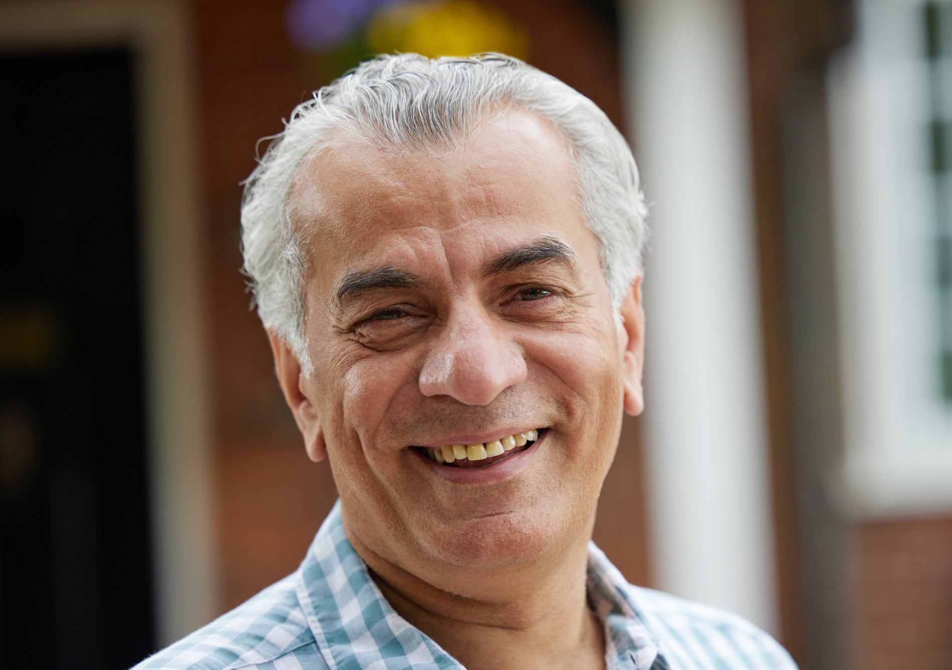 the photo shows an older man with dark eyes and grey hair standing outside and smiling at the camera